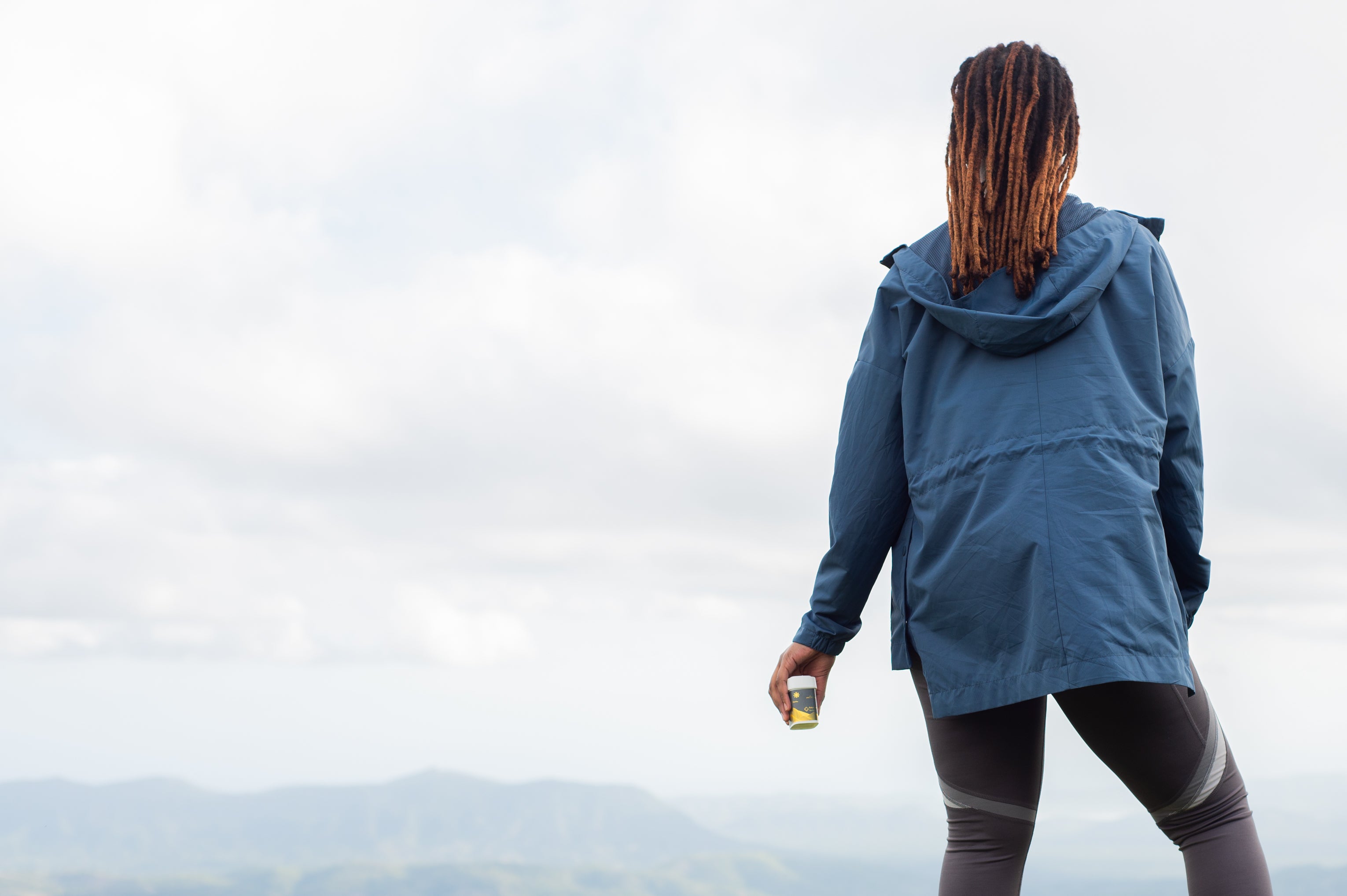 A person wearing a blue jacket is standing on a mountain with a cloudy sky, holding cannabis packaging.