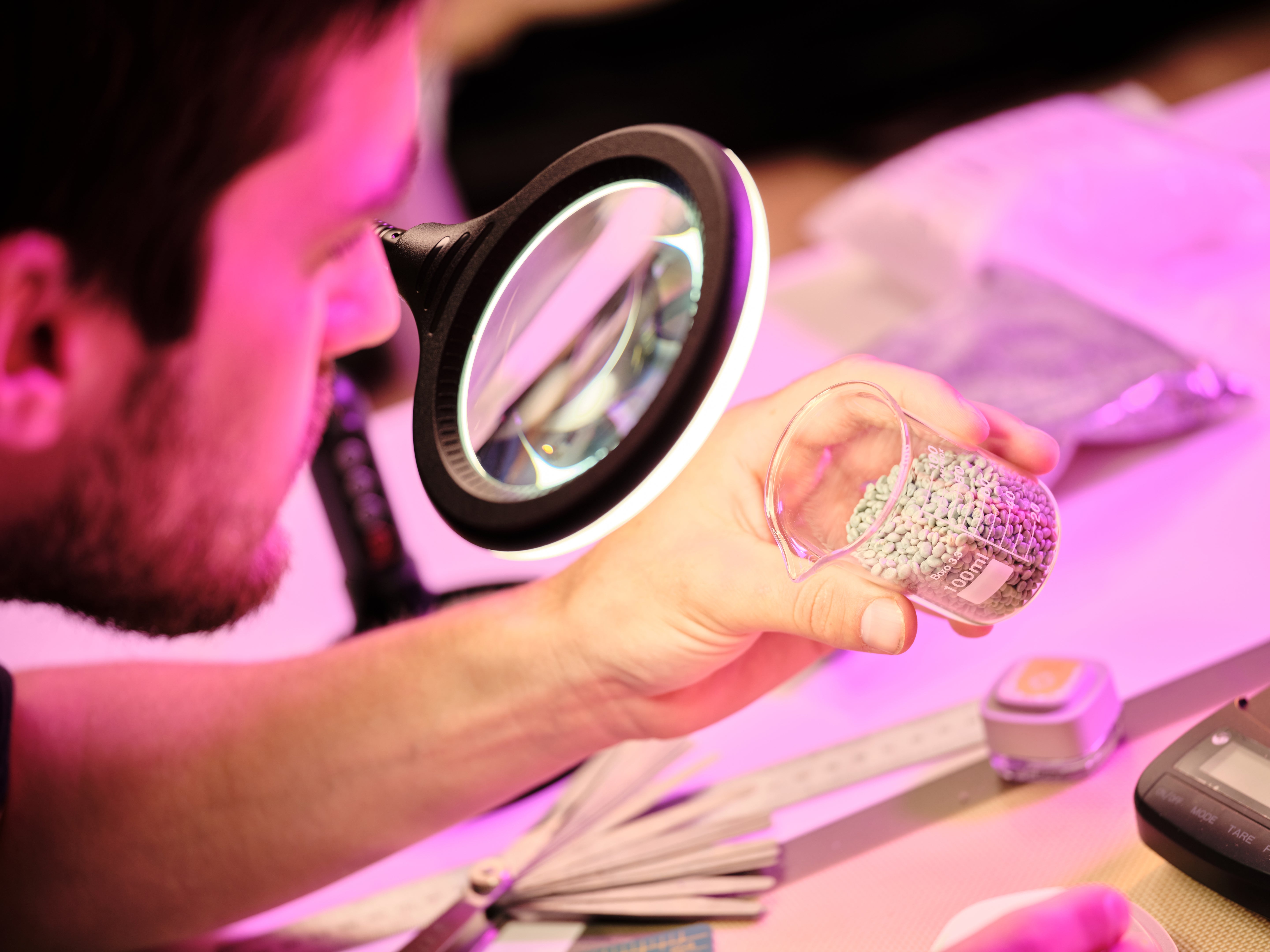 Person examining a small object under a magnifying glass with a pinkish-purple light.
